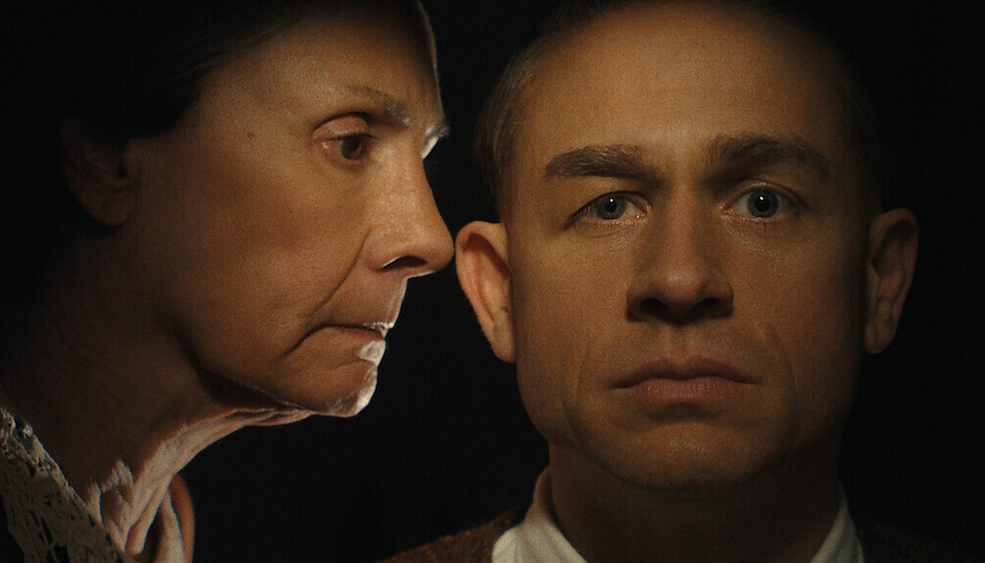 A cinematic image of an older woman on the verge of whispering something into a younger man's ear.