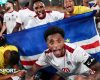 Cape Verde footballers celebrate a victory, with captain Ryan Mendes, wearing a white shirt with red trim, crouching down and extending his arms in front of a national flag and pulling a wide-eyed face of celebration with his mouth wide open