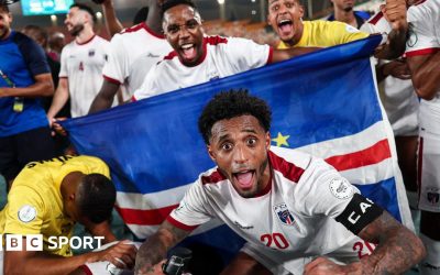 Cape Verde footballers celebrate a victory, with captain Ryan Mendes, wearing a white shirt with red trim, crouching down and extending his arms in front of a national flag and pulling a wide-eyed face of celebration with his mouth wide open