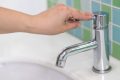 Imaginea, realizată în prim-plan, surprinde o mână care acționează maneta cromată a unei baterii moderne de chiuvetă, închizând robinetul. Deși robinetul este închis, o singură picătură de apă este vizibilă la capătul țevii, sugerând fie închiderea imediată, fie o scurgere minimă.