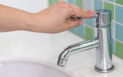 Imaginea, realizată în prim-plan, surprinde o mână care acționează maneta cromată a unei baterii moderne de chiuvetă, închizând robinetul. Deși robinetul este închis, o singură picătură de apă este vizibilă la capătul țevii, sugerând fie închiderea imediată, fie o scurgere minimă.