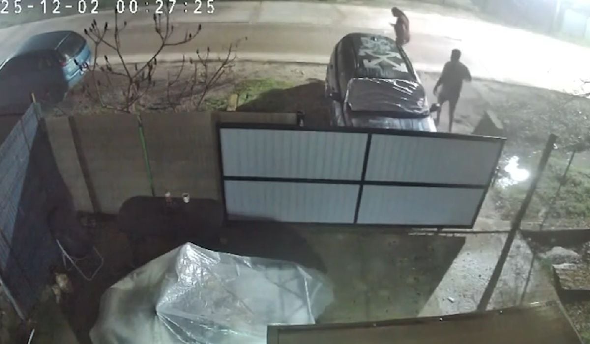Imagine surprinsă de o cameră de supraveghere, noaptea, arată două stând lângă o mașină parcată lângă un gard, pe marginea unui drum