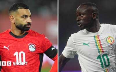 Mohamed Salah, wearing a red Egypt shirt with white 10 on the chest and a black captain's armband on his right bicep is seen looking to the right with his hands on his hips while Sadio Mane, wearing a white Senegal shirt with green, yellow and red detail and a green 10 on the chest, turns his head to the left